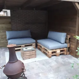 Zadaszone patio z dwoma sofami z palet z szarymi poduchami i poduszkami oparciowymi, drewnianym stolikiem kawowym i metalowym paleniskiem na pierwszym planie.