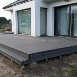 Nowoczesny taras z kompozytu w kolorze grafitowym, z dwoma stopniami, przylegający do białego budynku z dużymi oknami przesuwnymi i kwiatami w doniczce. W tle widoczny samochód dostawczy...