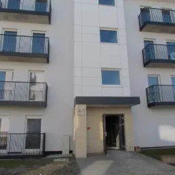 Nowoczesny budynek wielorodzinny z balkonami zabezpieczonymi czarnymi, metalowymi balustradami, widoczne wejście do budynku z numerem 44.
