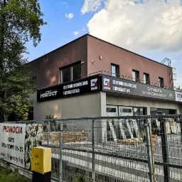Budynek centrum budowlanego i brukarstwa z szyldami reklamowymi, widok z zewnątrz przez metalowe ogrodzenie, na pierwszym planie baner z promocją grysu granitowego.