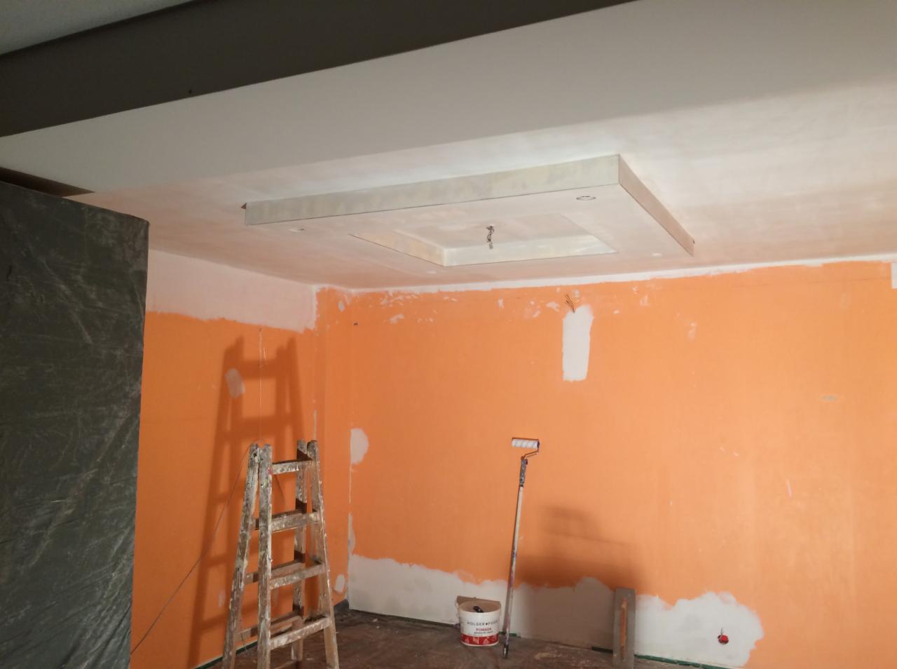 Pokój w trakcie malowania na pomarańczowo, widoczna drabina, wałek malarski oparty o ścianę i wiadro z farbą, sufit z dekoracyjnym podwieszanym elementem.