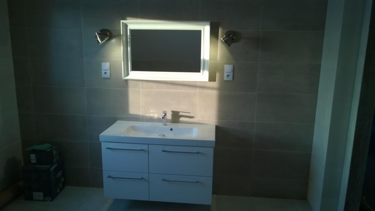 Wykończona łazienka z szarymi płytkami, białą szafką pod umywalką i lustrem w białej ramie, oświetlona dwoma metalowymi lampami punktowymi.