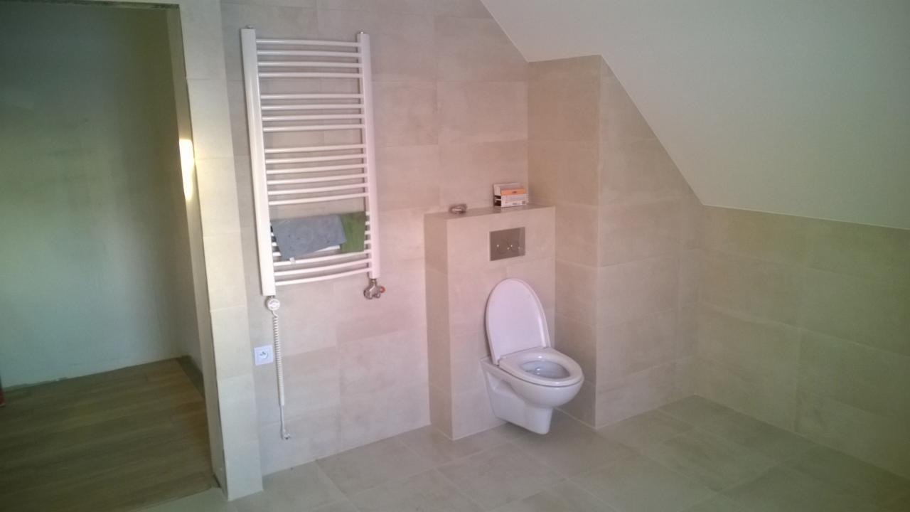 Wykończona łazienka w odcieniach beżu z podwieszaną toaletą, grzejnikiem drabinkowym z ręcznikiem i widocznym wejściem do drugiego pomieszczenia w stanie surowym.