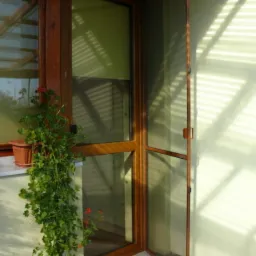 Otwarta drewniana moskitiera drzwiowa z drobną siatką, widoczna z bliska, z pelargonią w doniczce na parapecie okna w tle.