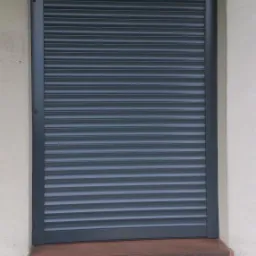 Zamknięta, szara osłona okienna z lamelami, zamontowana na zewnątrz budynku z widocznym parapetem.