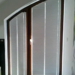 Dwuskrzydłowe okno o nietypowym kształcie z zamontowanymi białymi plisami, widoczne promienie słoneczne na podłodze.