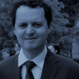 Portret uśmiechniętego mężczyzny w ciemnym garniturze i krawacie w kropki, z zamazanym tłem parkowym.