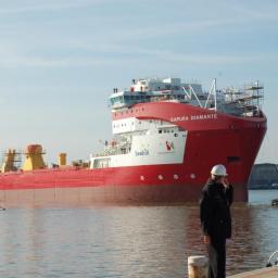 NUEVA TERRAIN POLSKA SP. Z O.O. - Czerwono-biały statek Sapura Diamante na wodzie, z mężczyzną w kasku rozmawiającym przez telefon na pierwszym planie.