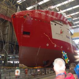 NUEVA TERRAIN POLSKA SP. Z O.O. - Czerwono-biały statek 'SAPURA DIAMANTE' w doku, udekorowany balonami i konfetti podczas ceremonii wodowania, widok z perspektywy obserwatorów.