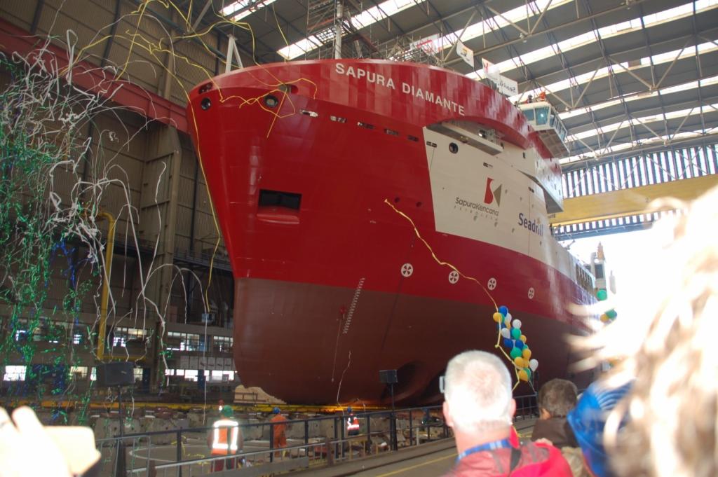 Czerwono-biały statek 'SAPURA DIAMANTE' w doku, udekorowany balonami i konfetti podczas ceremonii wodowania, widok z perspektywy obserwatorów.