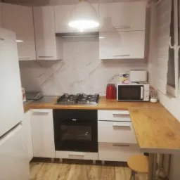 Nowoczesna biała kuchnia z drewnianym blatem, marmurowym backsplashiem i wiszącą lampą, z widocznym sprzętem AGD: lodówką, piekarnikiem, płytą gazową i mikrofalówką.