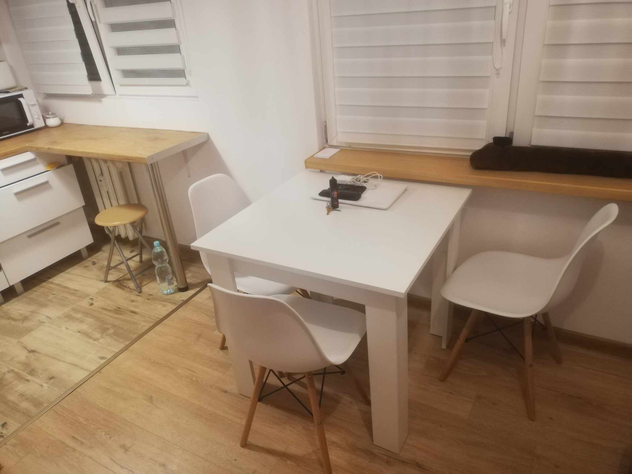 Jasna kuchnia z drewnianą podłogą, białymi meblami, stołem i krzesłami w stylu skandynawskim, z laptopem i akcesoriami na blacie stołu.