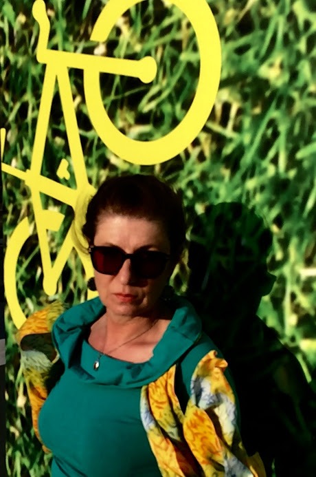 Kobieta w okularach przeciwsłonecznych pozuje na tle zielonej ściany z żółtym symbolem roweru.