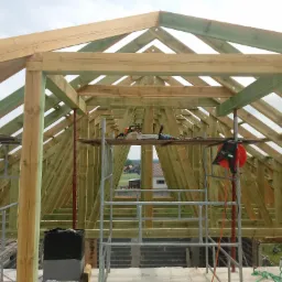 Drewniana konstrukcja dachu w trakcie budowy, widoczne narzędzia na rusztowaniu i elementy konstrukcyjne pomalowane na zielono.