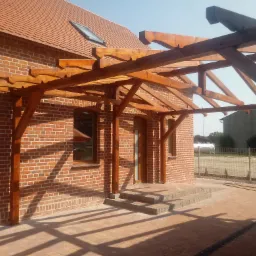 Nowoczesna pergola drewniana zadaszająca wejście do budynku z czerwonej cegły, widoczne schody wejściowe i dachówka ceramiczna.