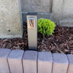 Szara kolumna z panelem sterowania Nice przy bramie, obok betonowy słupek i mała tuja, kora na ziemi, obrzeże z kostki.