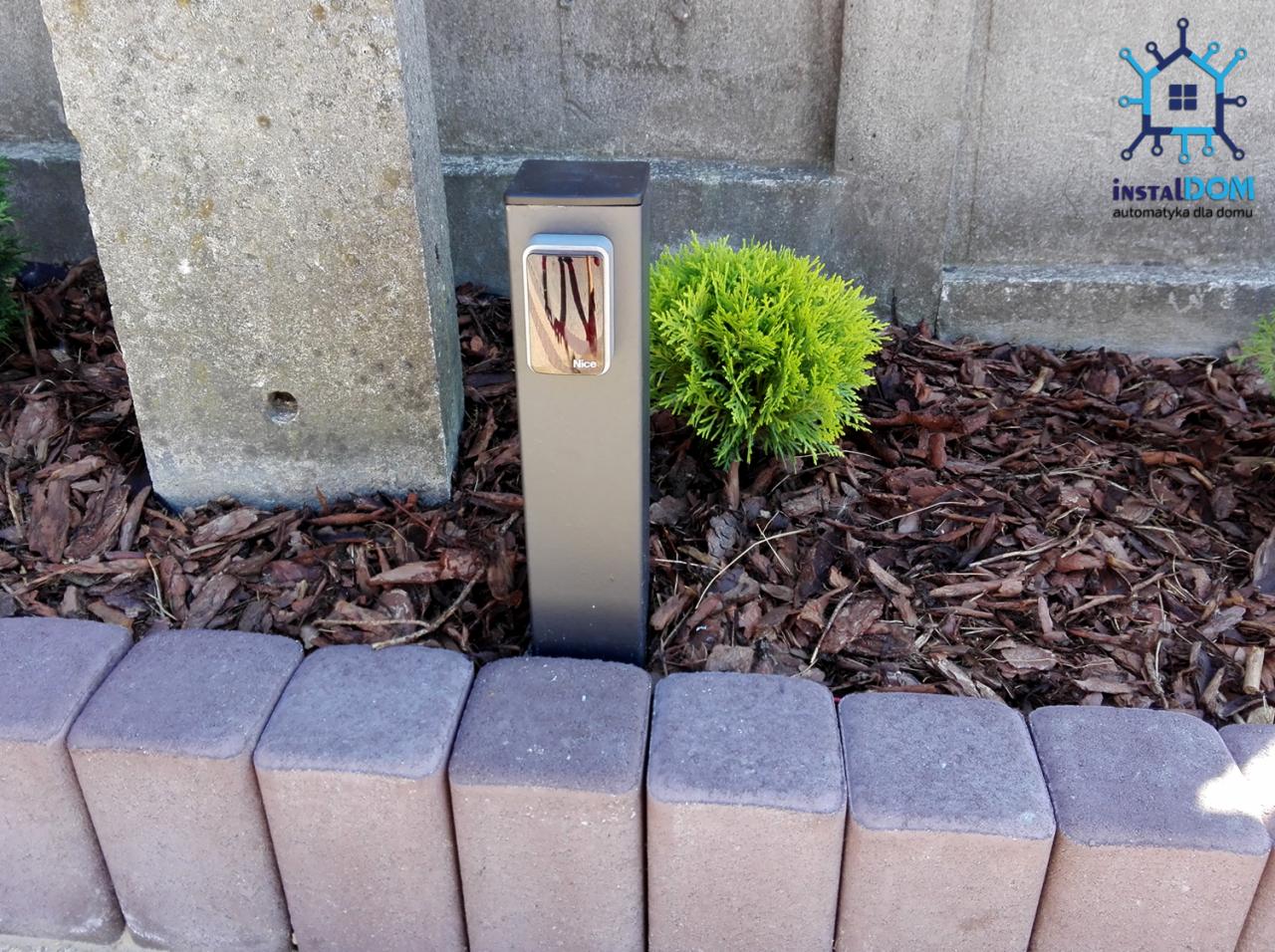 Szara kolumna z panelem sterowania Nice przy bramie, obok betonowy słupek i mała tuja, kora na ziemi, obrzeże z kostki.