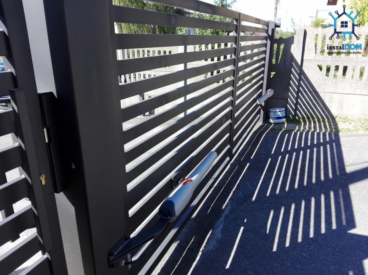Nowoczesna, czarna, przesuwna brama z automatyką, widoczne siłowniki, cień bramy na asfalcie, w tle ogrodzenie betonowe i zieleń.