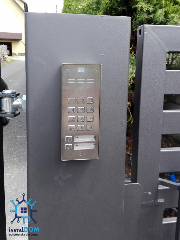 Szary panel z klawiaturą numeryczną i dwoma przyciskami dzwonka zamontowany na słupku bramy wjazdowej, widoczny fragment bramy i logo firmy instalacyjnej w lewym dolnym rogu.