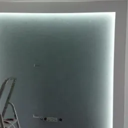 Podświetlona ściana z efektem dekoracyjnym w ramie, widoczna drabina malarska, gniazdka elektryczne w dolnej części ściany, fragment sufitu i ściany w tle.