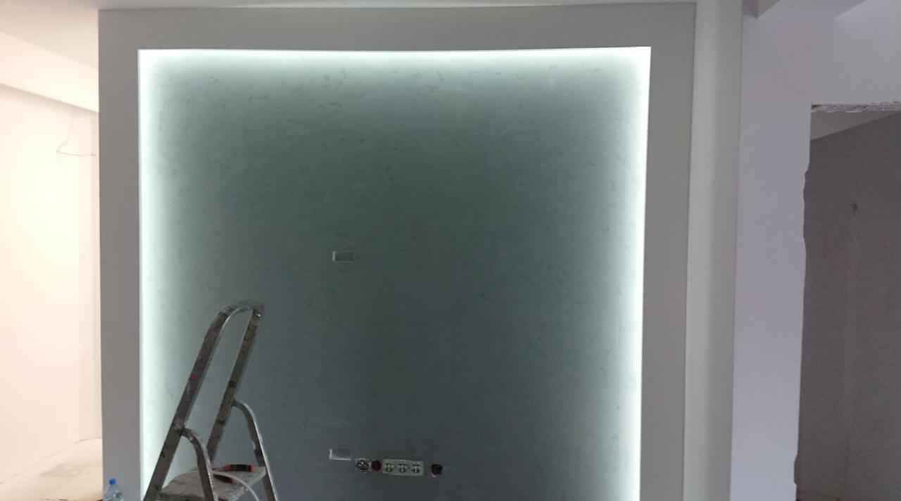 Podświetlona ściana z efektem dekoracyjnym w ramie, widoczna drabina malarska, gniazdka elektryczne w dolnej części ściany, fragment sufitu i ściany w tle.