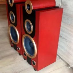 Para czerwonych kolumn głośnikowych z czarnymi panelami i złotymi obwódkami na drewnianej podłodze w pomieszczeniu z szarą szafką i dekoracyjną ścianą w tle.