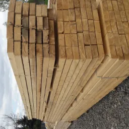 Stos tarcicy budowlanej ułożony na żwirowym podłożu, widoczne końcówki desek z wyraźnymi słojami drewna, fragment pochmurnego nieba w tle.