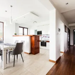 Jasne, przestronne wnętrze domu: jadalnia z białym stołem i szarymi krzesłami, otwarta na kuchnię z drewnianym blatem i białą zabudową, oraz korytarz z ciemną drewnianą podłogą i schodami.