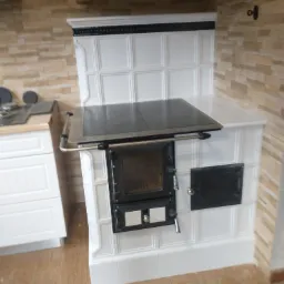 Biały piec kaflowy z czarnymi elementami, z blatem i drzwiczkami, ustawiony w rogu kuchni z drewnianą podłogą i ścianą wyłożoną kamieniem dekoracyjnym.