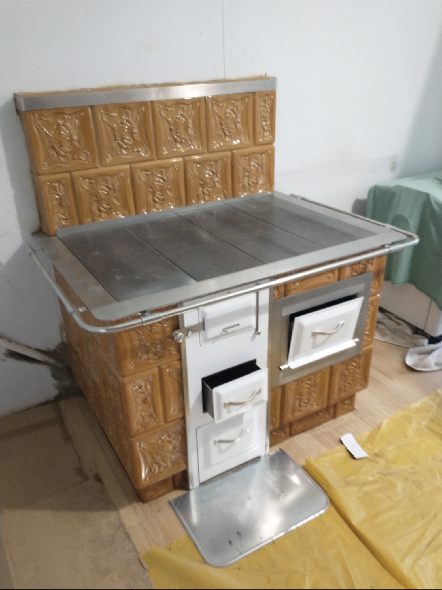 Nowoczesny piec kaflowy z beżowymi kaflami z wytłaczanym wzorem, z metalowym blatem i białymi drzwiczkami szuflad, na tle białej ściany i podłogi zabezpieczonej folią.