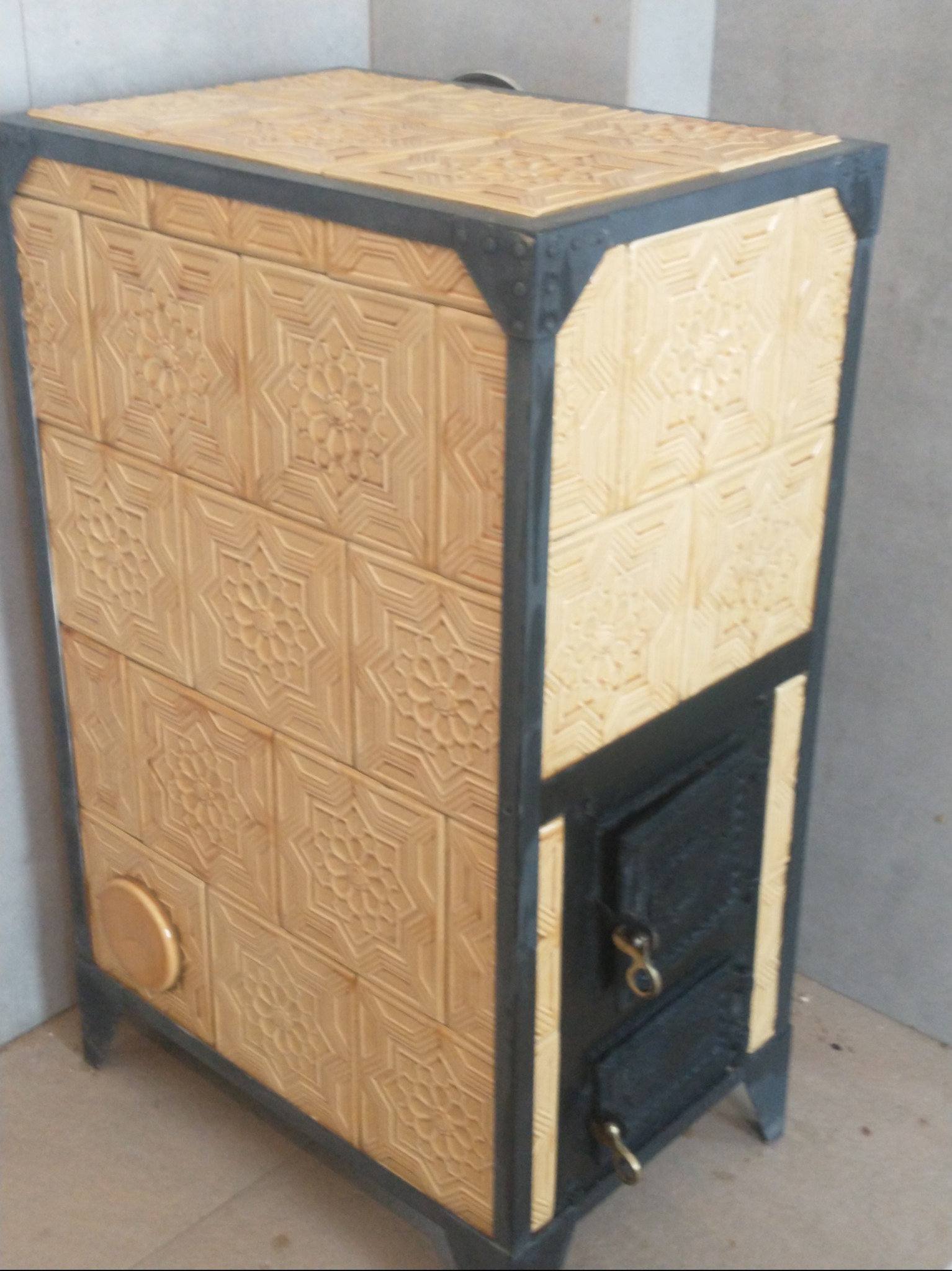 Kafelkowy piec z dekoracyjnymi wzorami kwiatowymi, obudowany metalową ramą z czarnymi drzwiczkami paleniska i popielnika.