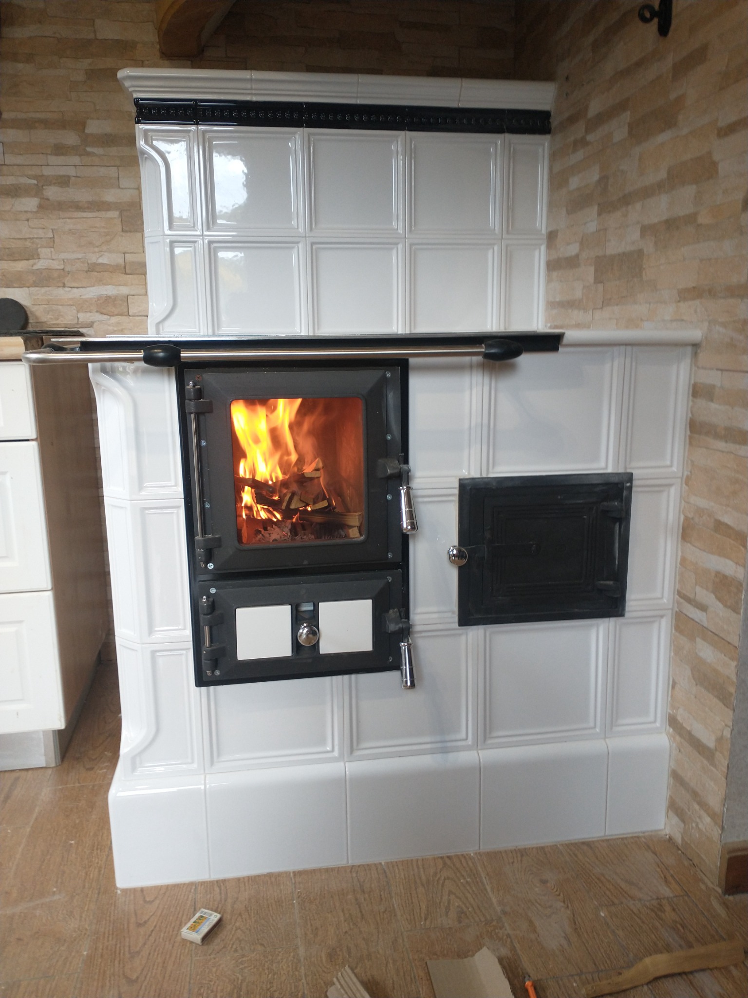 Biały piec kaflowy z czarnymi drzwiczkami, w których widać płomienie. Obok pieca widoczna ściana z kamienia dekoracyjnego i fragment podłogi z pudełkiem zapałek.