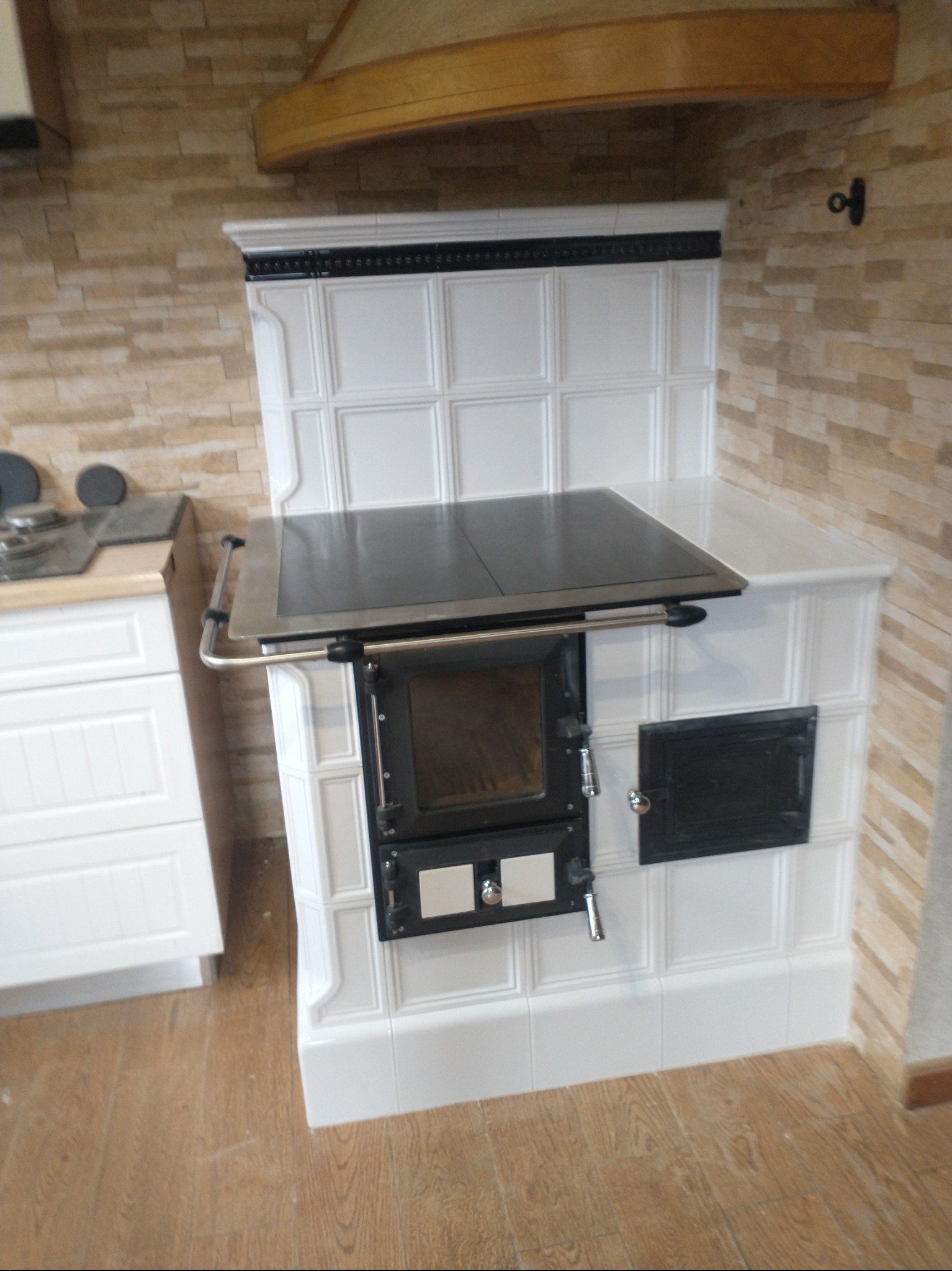 Biały piec kaflowy z czarnymi elementami, z blatem i drzwiczkami, ustawiony w rogu kuchni z drewnianą podłogą i ścianą wyłożoną kamieniem dekoracyjnym.