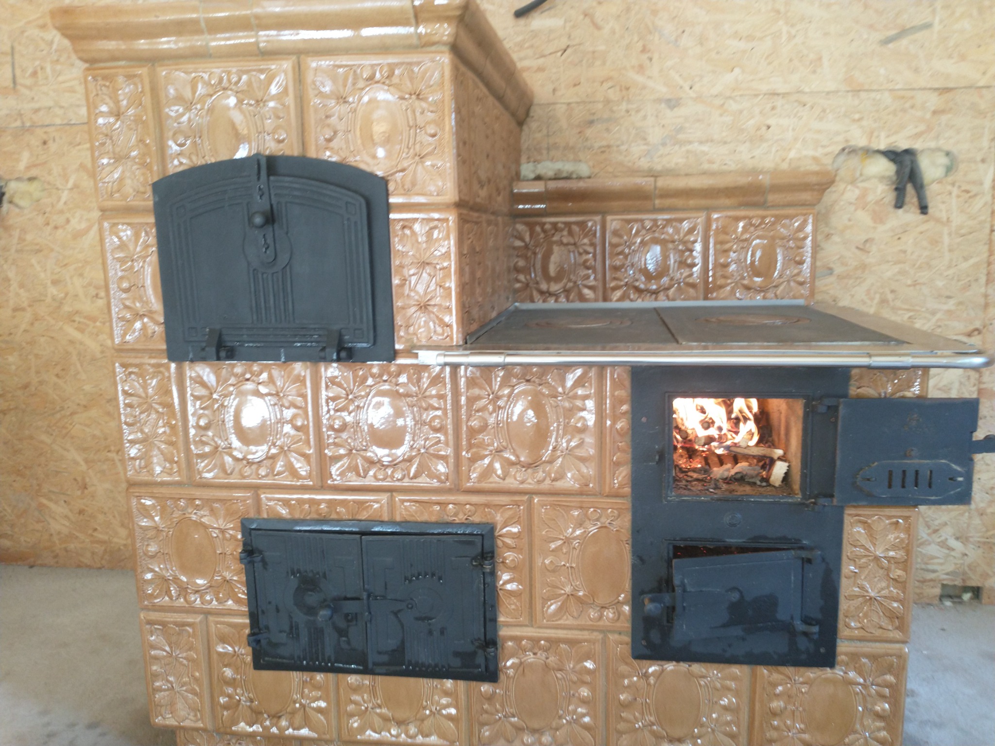 Kuchenka kaflowa z otwartymi drzwiczkami paleniska, w którym widać płomienie, oraz z metalowym blatem nad paleniskiem, obudowana płytkami ceramicznymi w odcieniach beżu z tłoczonym wzorem.