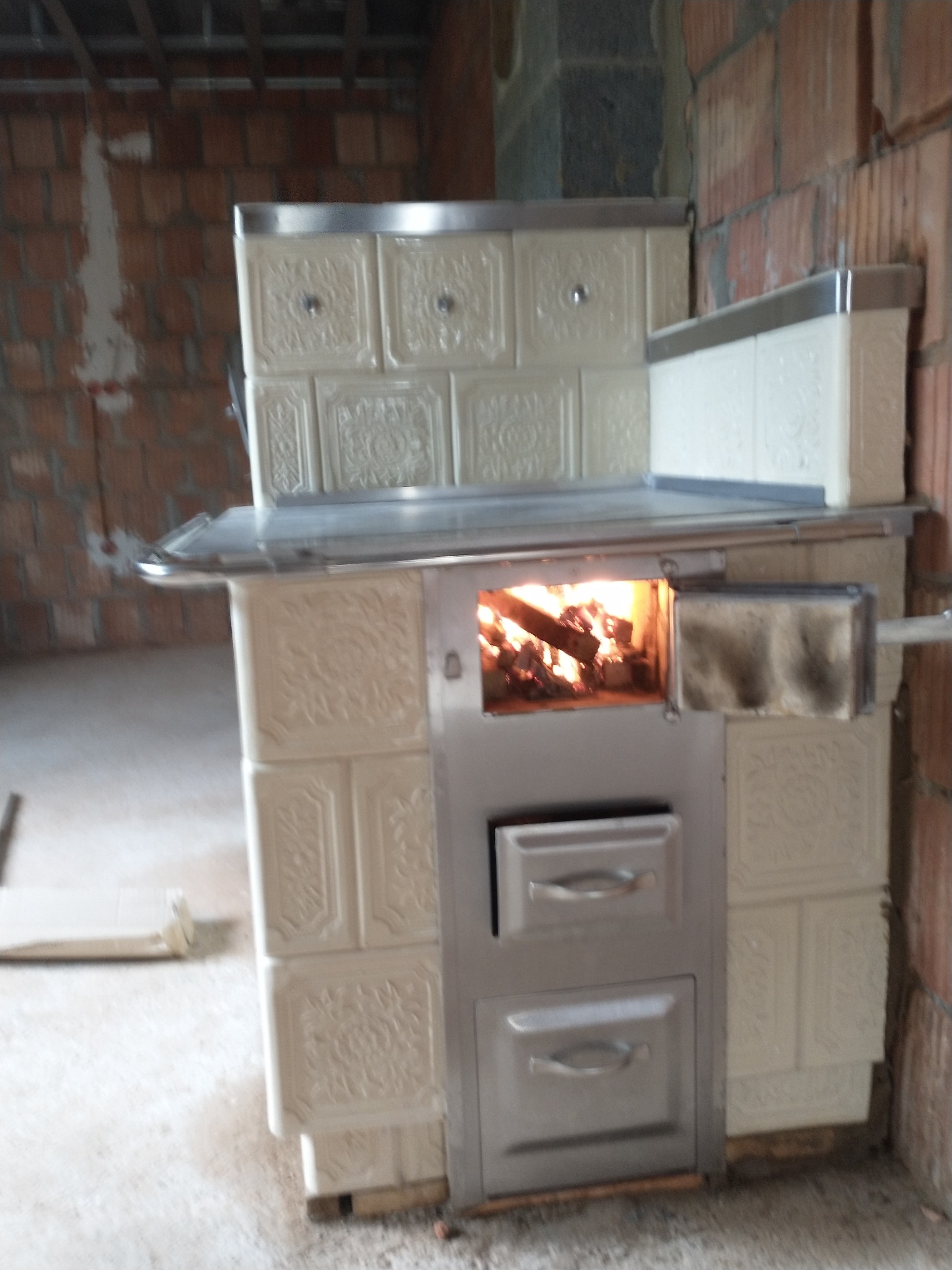 Wnętrze w trakcie budowy z widocznym piecem kaflowym z paleniskiem, w którym płonie ogień, oraz dwoma metalowymi drzwiczkami poniżej.