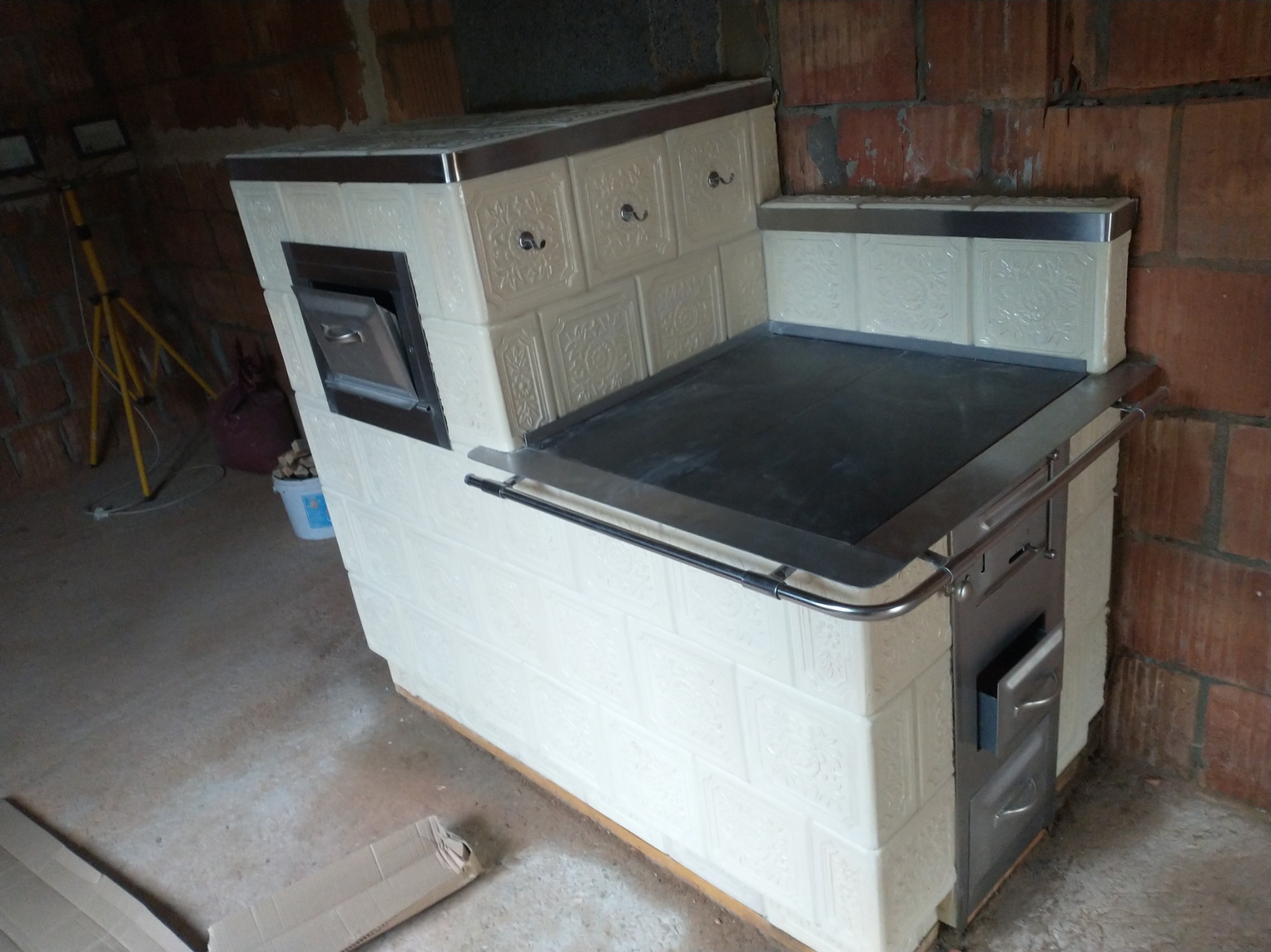 Biały piec kaflowy z czarną płytą grzewczą i metalowymi elementami wykończeniowymi, w surowym wnętrzu z ceglanymi ścianami.