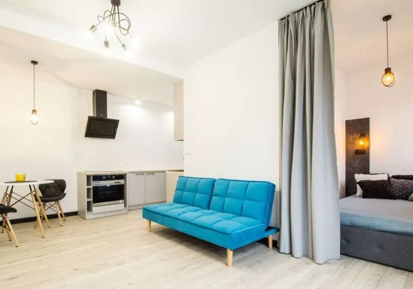 Małe mieszkanie typu studio z aneksem kuchennym, przedzielone zasłoną, z turkusową sofą na pierwszym planie i minimalistycznym oświetleniem.