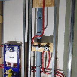 Instal P&P - Wewnętrzna instalacja wodno-kanalizacyjna w trakcie montażu, widoczne rury szare i czerwone, stelaż podtynkowy Geberit, konstrukcja z profili stalowych, surowy stan wykończenia.