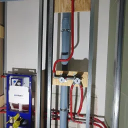 Wewnętrzna instalacja wodno-kanalizacyjna w trakcie montażu, widoczne rury szare i czerwone, stelaż podtynkowy Geberit, konstrukcja z profili stalowych, surowy stan wykończenia.