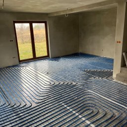 Instal P&P - Niebieskie rury ogrzewania podłogowego ułożone na folii w surowym wnętrzu domu w trakcie budowy, widok na okno i ogród.