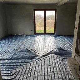 Instal P&P - Niedokończona instalacja ogrzewania podłogowego z niebieskimi rurami na folii w surowym, jasnym pomieszczeniu z dużym oknem i widokiem na drzewa.