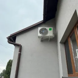 Zewnętrzna jednostka klimatyzacji Kaisai zamontowana na szarej elewacji domu, widoczny brązowy system rynnowy i okno z drewnianą ramą w pochmurny dzień.