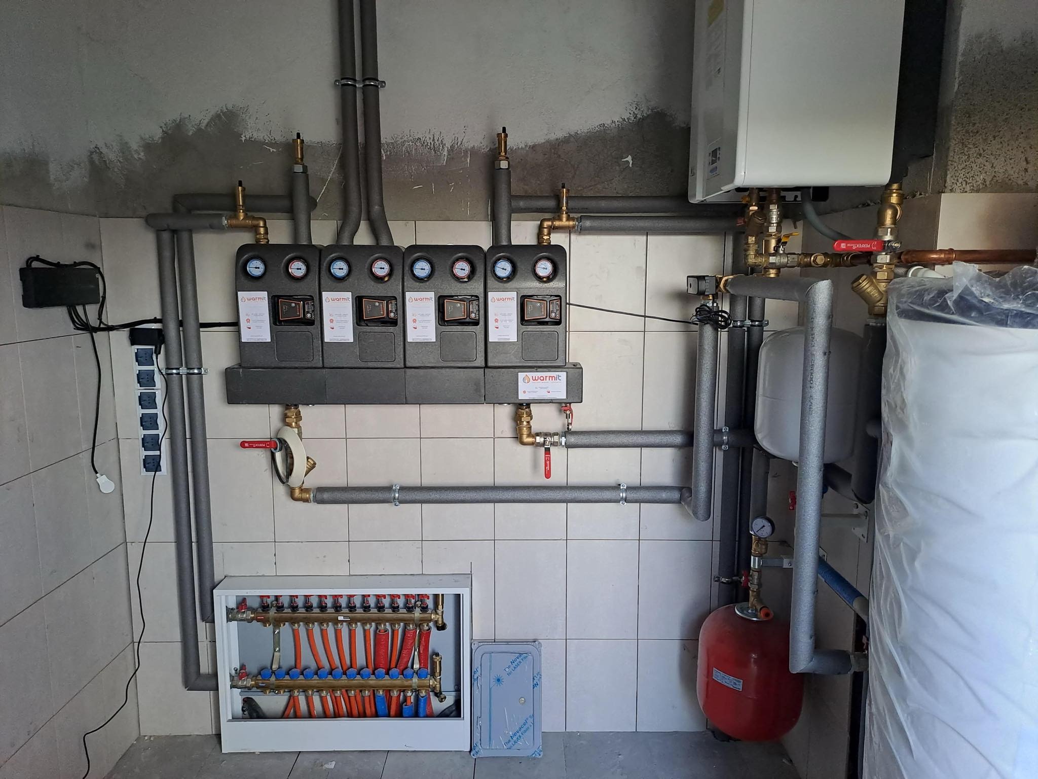 Rozdzielacz ogrzewania podłogowego z czterema modułami sterującymi Warmit, zbiornikiem wyrównawczym i piecem w surowym pomieszczeniu z kafelkami na ścianach.