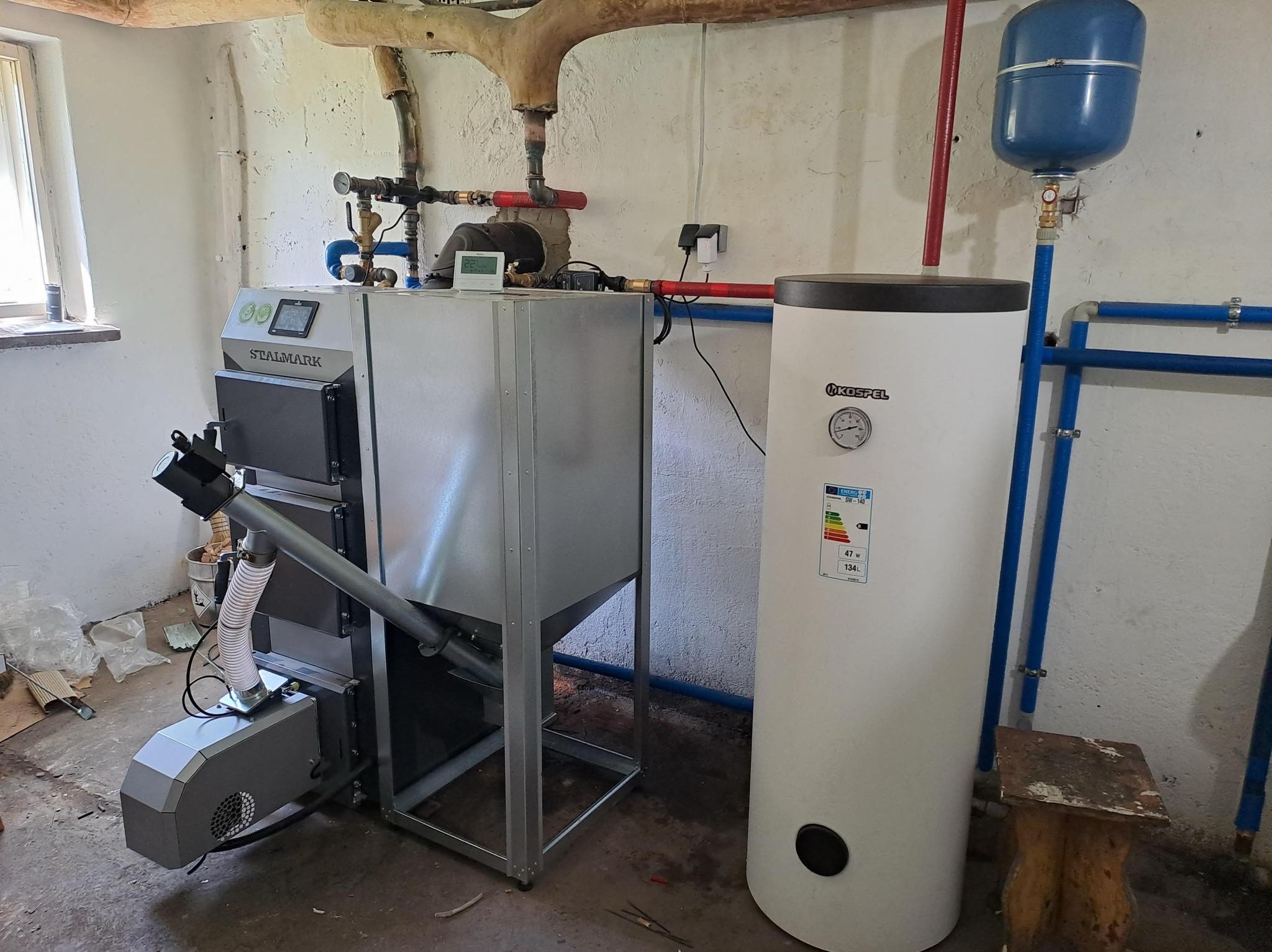 Kotłownia z piecem na paliwo stałe Stalmark z podajnikiem ślimakowym, zasobnikiem ciepłej wody Kospel i instalacją hydrauliczną w surowym pomieszczeniu.