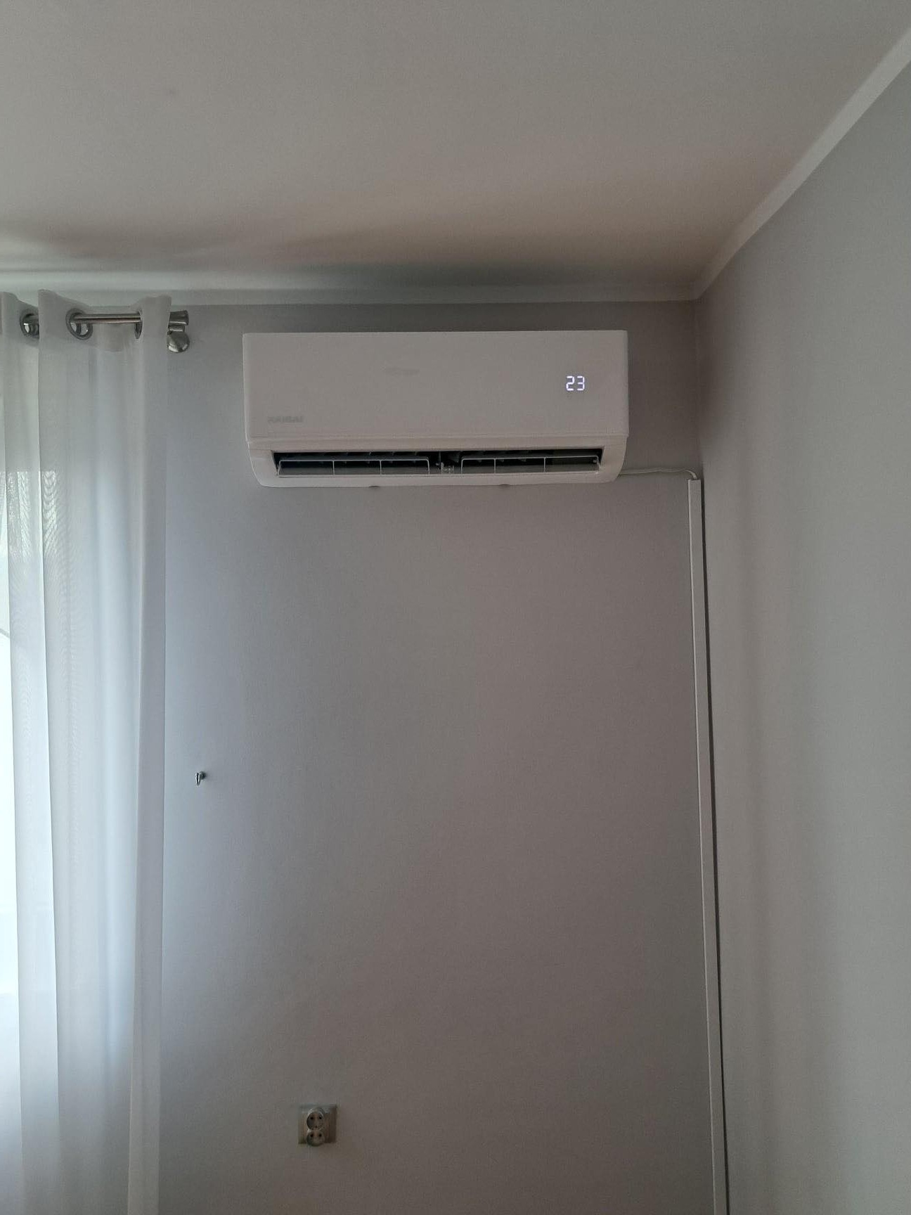 Biały klimatyzator ścienny z wyświetlaczem temperatury 23 stopnie Celsjusza, zamontowany na szarej ścianie obok zwiewnej, białej zasłony.