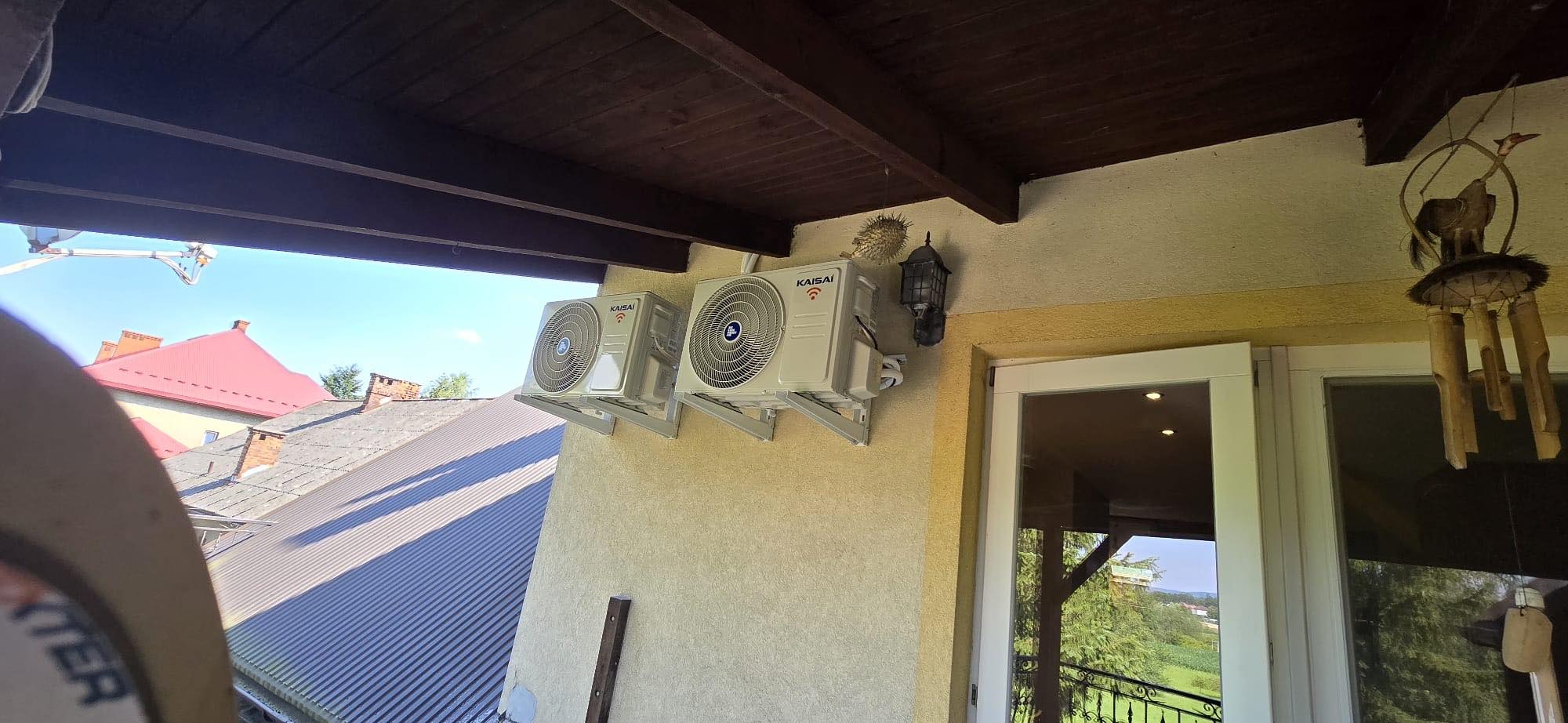 Dwa klimatyzatory marki Kaisai zamontowane na elewacji budynku z widokiem na dachy sąsiednich domów i dekoracyjną latarnię.