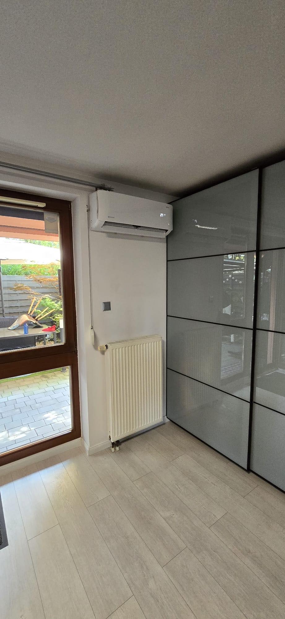 Biały klimatyzator ścienny z wyświetlaczem temperatury zamontowany nad grzejnikiem w jasnym pomieszczeniu z drewnianą podłogą i przesuwnymi drzwiami szafy.