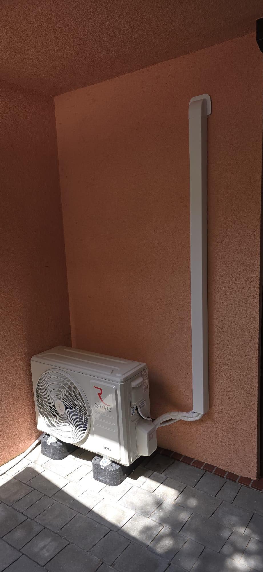 Zewnętrzna jednostka klimatyzacji Rotenso zamontowana na gumowych podkładkach na zewnątrz budynku, z widoczną maskownicą przewodów na ścianie.