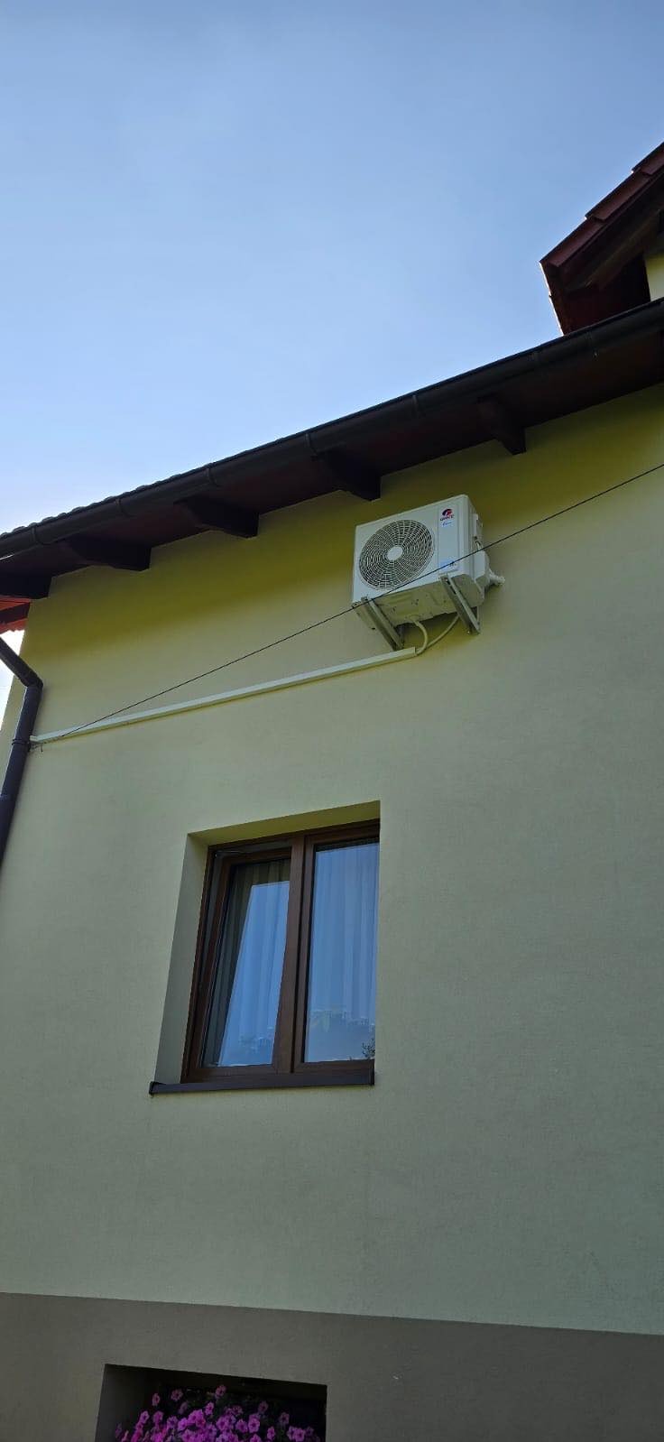 Zewnętrzna jednostka klimatyzacji zamontowana pod okapem budynku o kremowej elewacji, widoczna instalacja elektryczna, okno z brązowymi ramami i zasłoną.