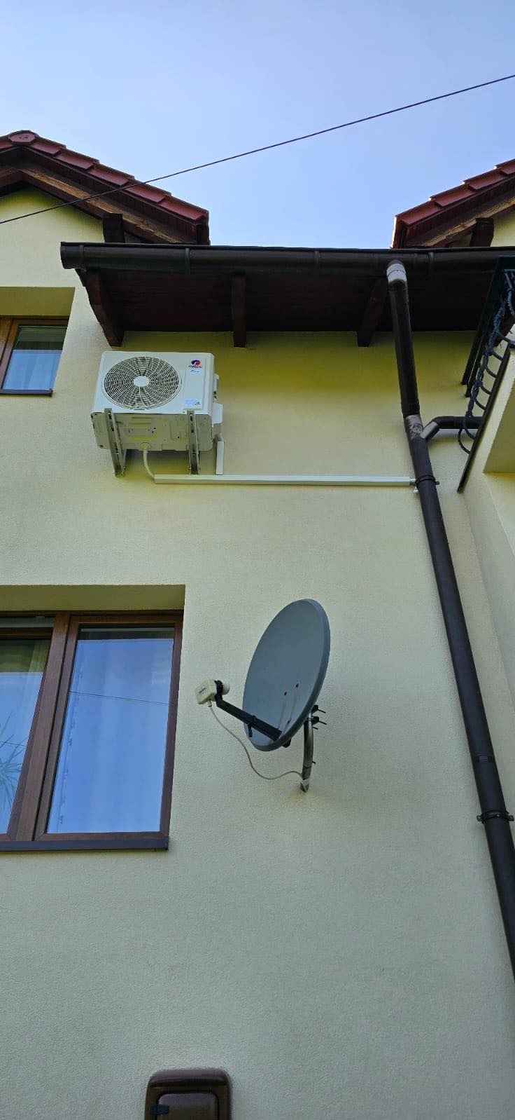 Zewnętrzna jednostka klimatyzacji zamontowana na elewacji domu obok okna i rynny, widoczna także antena satelitarna.